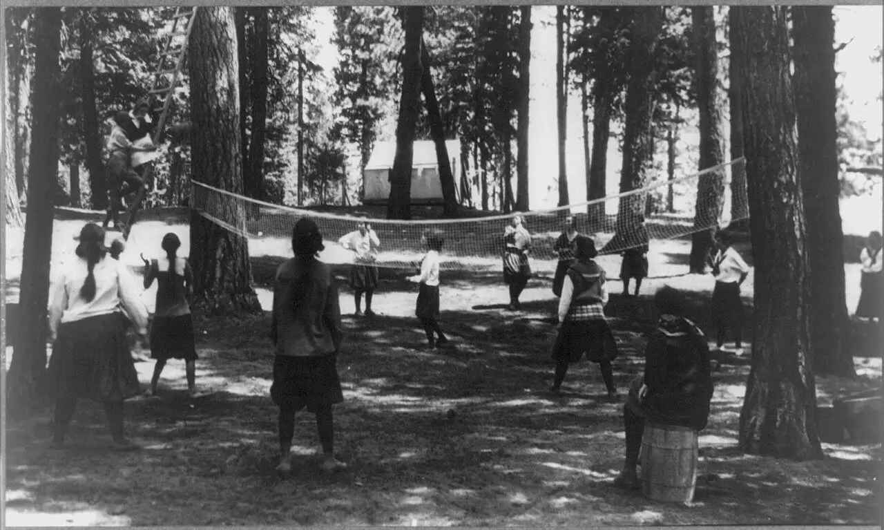 Young women playing volleyball among tall pine trees at a YWCA camp at Payette Lake, Idaho, circa 1912-1920