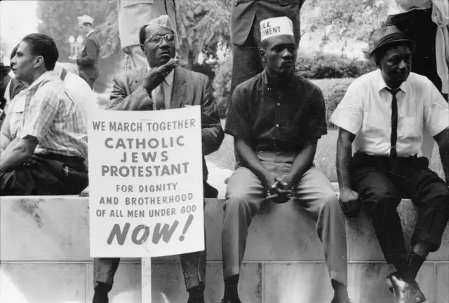 Black-and-white 1965 photograph of civil rights marchers resting with a sign reading 'We march together — Catholic, Jews, Protestant — for dignity and brotherhood of all men under God, now!'