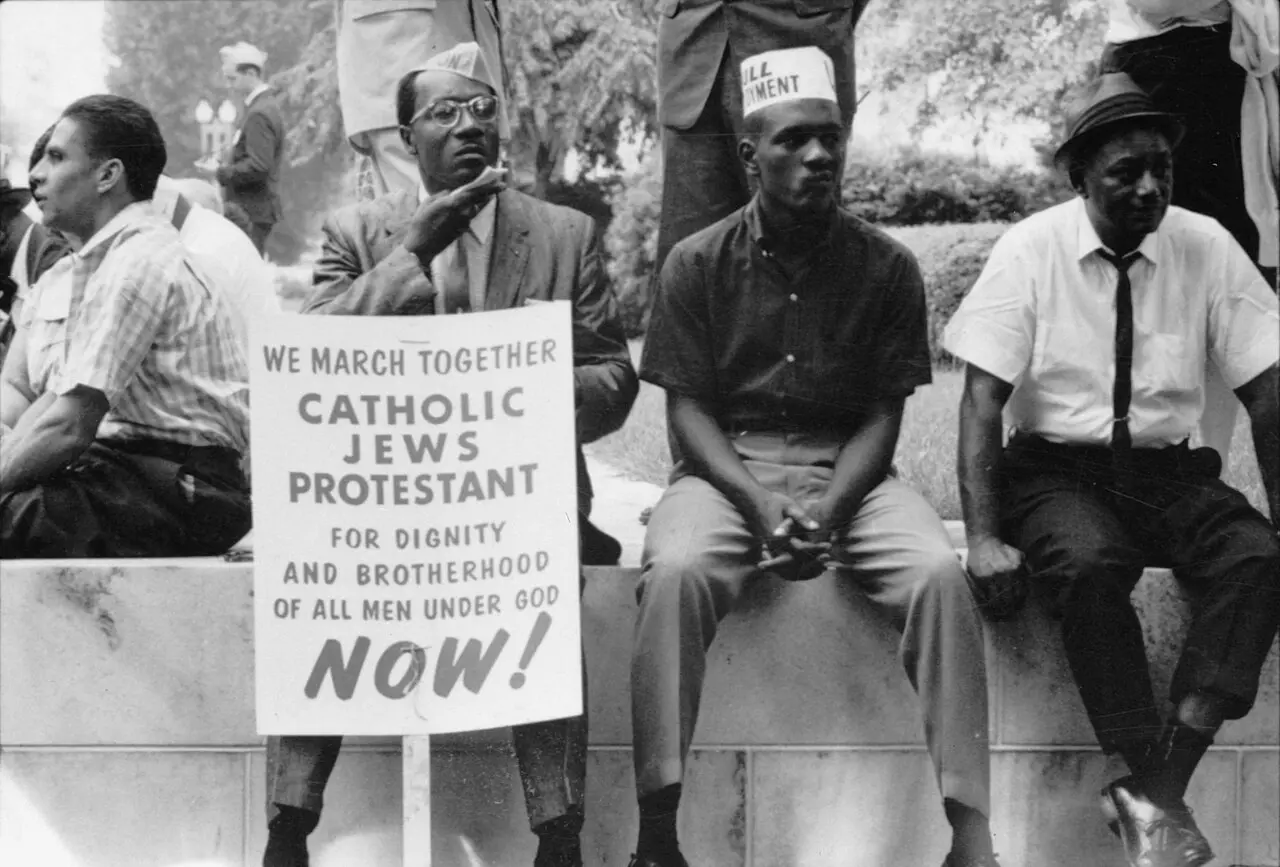 Black-and-white 1965 photograph of civil rights marchers resting with a sign reading 'We march together — Catholic, Jews, Protestant — for dignity and brotherhood of all men under God, now!'