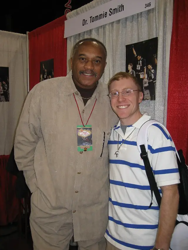 Tommie Smith photographed at a 2007 marathon expo, standing beside a display showing the famous 1968 Olympic podium protest with raised fists.