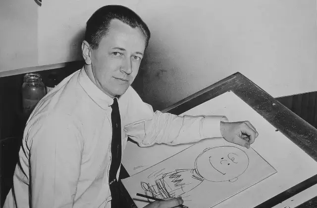 Black-and-white 1956 photograph of Peanuts creator Charles Schulz sketching Charlie Brown at his drawing board.