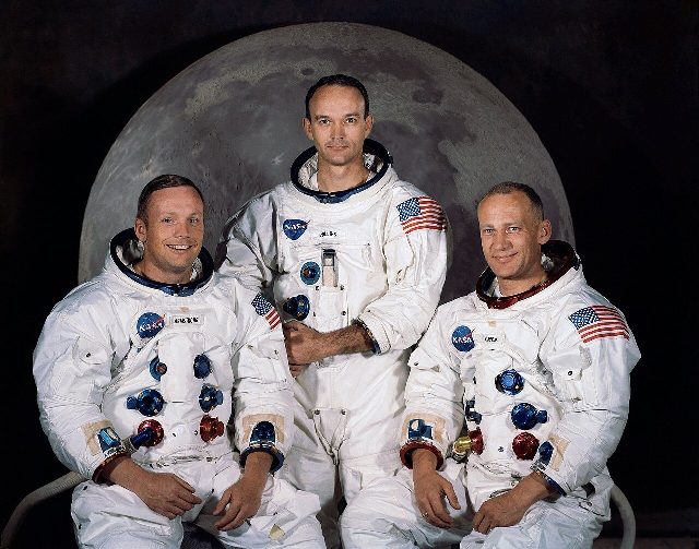 The Apollo 11 crew — Neil Armstrong, Michael Collins, and Buzz Aldrin — in their spacesuits against a backdrop of the Moon.