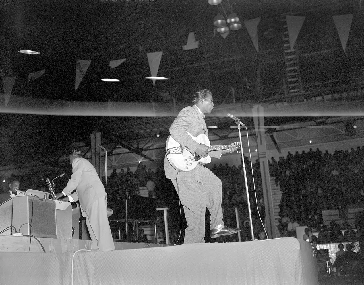 Chuck Berry duck-walking across a stage with his guitar at a 1957 concert in Edmonton, Alberta, before a packed crowd.
