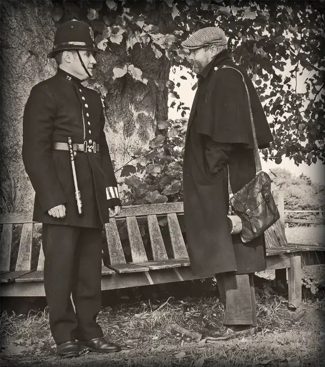 A reenactor dressed as a Victorian-era London police constable in a top hat and long blue coat, speaking with a costumed civilian
