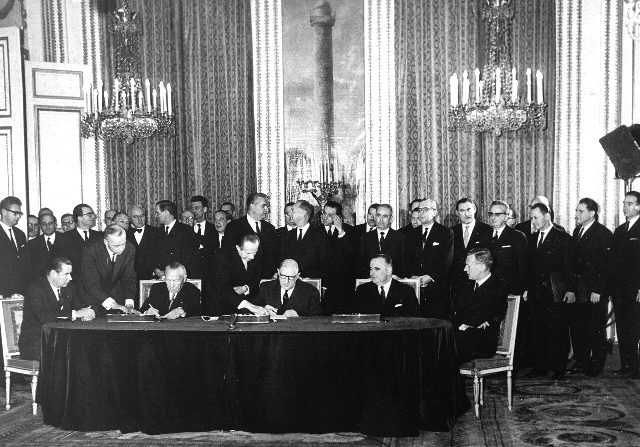 A black-and-white 1963 photograph of the Élysée Treaty signing ceremony in a grand chandeliered hall of the Élysée Palace, with de Gaulle and Adenauer seated at a long table surrounded by officials.