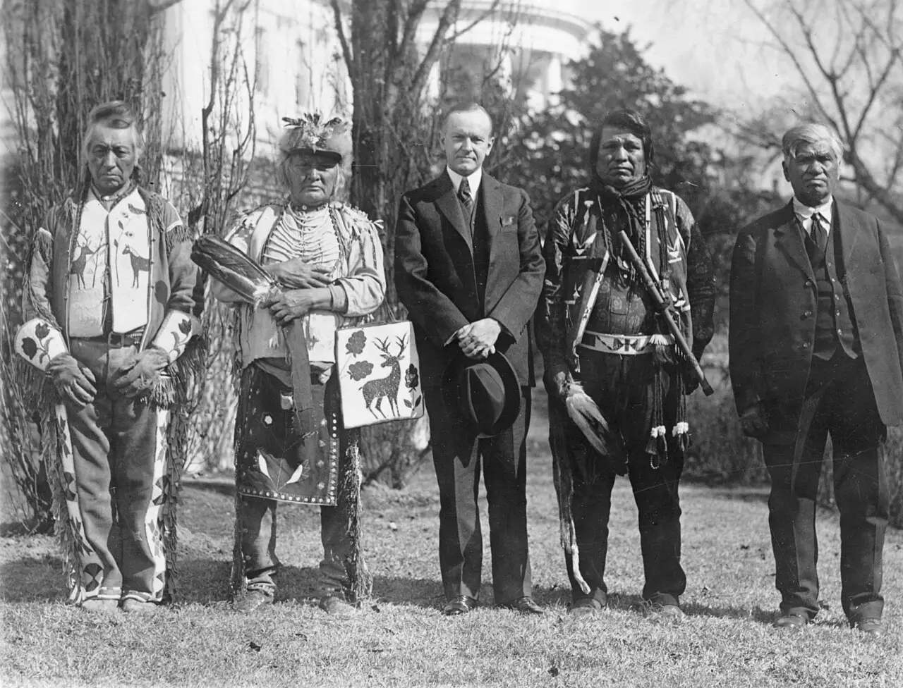 Black-and-white photograph of President Calvin Coolidge standing with four Osage Indians in traditional dress on the White House grounds.