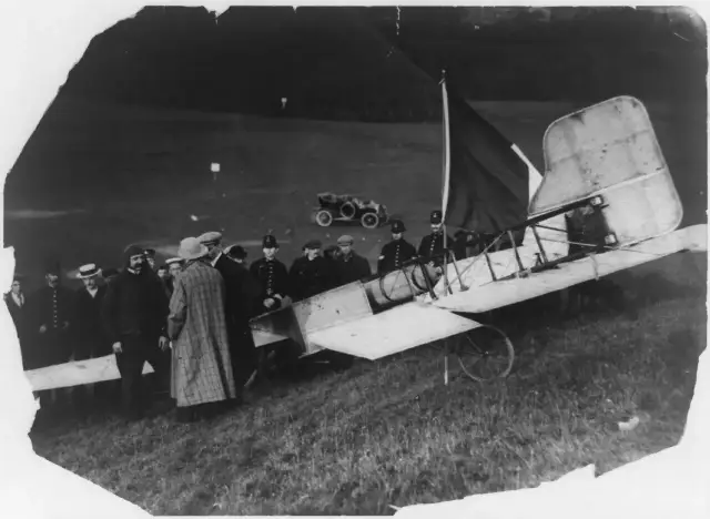 Louis Blériot standing beside his damaged Blériot XI monoplane with a French tricolor flag, surrounded by onlookers after landing near Dover on July 25, 1909