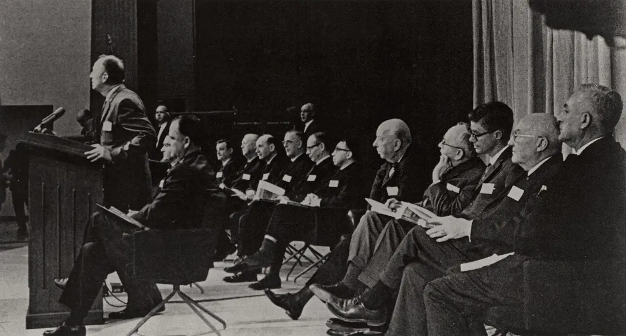 Surgeon General Luther Terry speaking at a podium during the January 11, 1964 press conference releasing the report on smoking and health, with committee members seated behind him.
