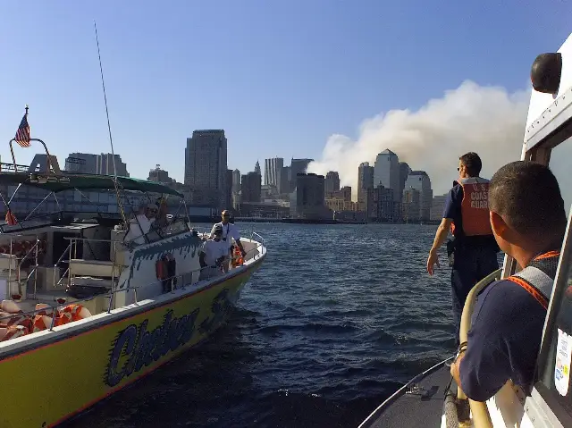 A U.S. Coast Guard boarding team in an orange life vest aboard a boat in New York Harbor on September 11, 2001, with smoke from the World Trade Center site rising in the background.