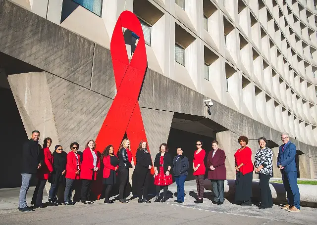 People in red clothing gathered in front of a giant red AIDS awareness ribbon displayed on a government building for World AIDS Day