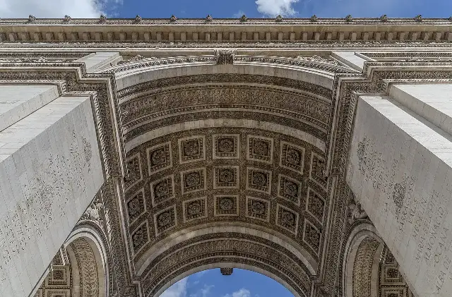 Looking up through the vaulted arch of the Arc de Triomphe in Paris, with the names of Napoleonic battlefields carved into the stone overhead.