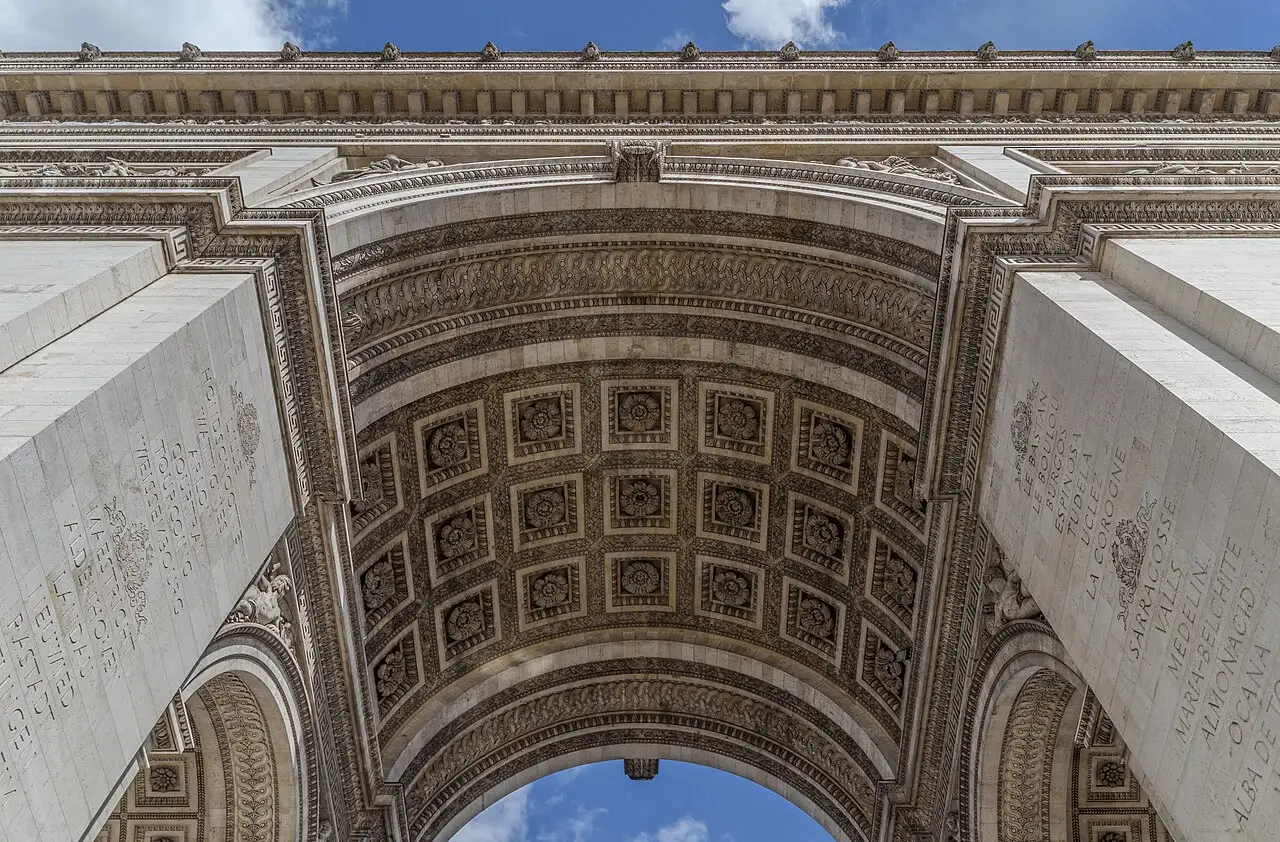 Looking up through the vaulted arch of the Arc de Triomphe in Paris, with the names of Napoleonic battlefields carved into the stone overhead.