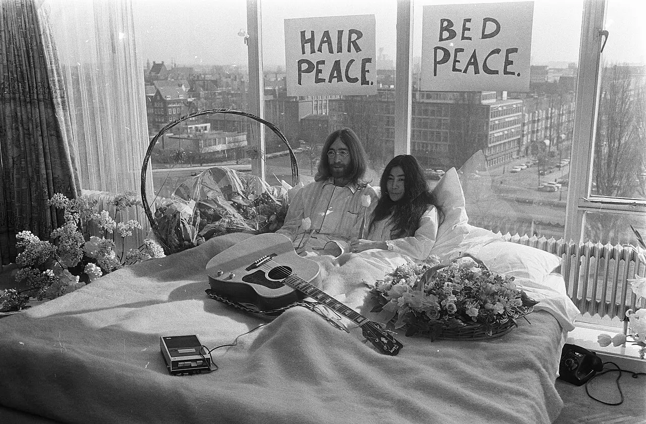 John Lennon and Yoko Ono during their 1969 bed-in for peace in Amsterdam, behind hand-lettered signs reading 'Hair Peace' and 'Bed Peace'.