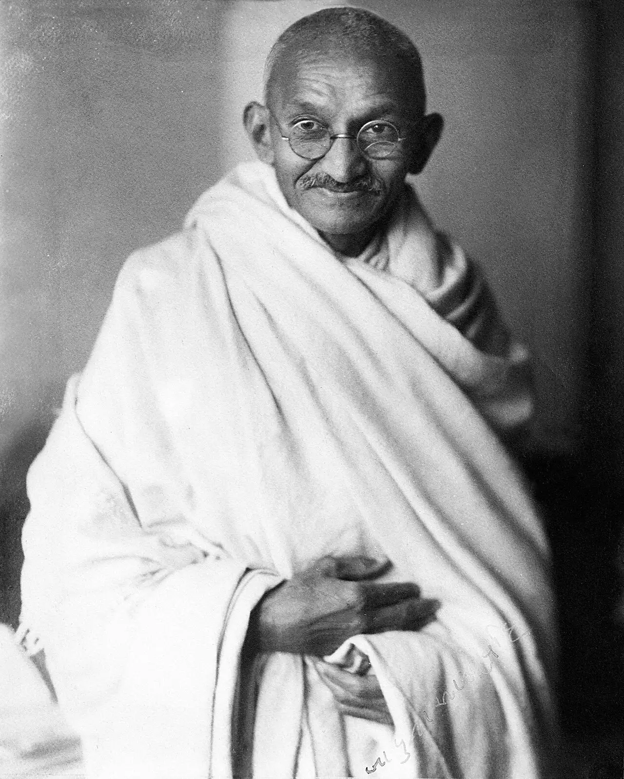 Black-and-white 1931 studio portrait of Mahatma Gandhi wearing a white dhoti and shawl, smiling gently at the camera