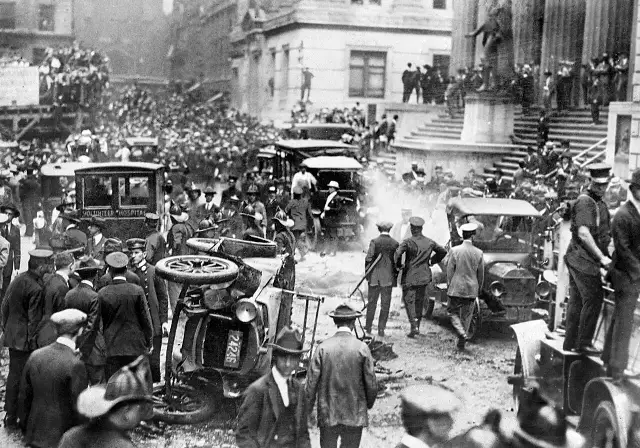 Black-and-white 1920 photograph of the Wall Street bombing aftermath: a wrecked car, a Volunteer Hospital ambulance, and crowds outside 23 Wall Street