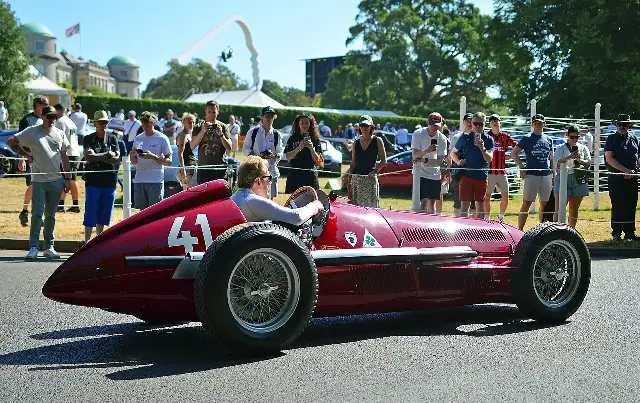 A red 1950 Alfa Romeo 158 'Alfetta' grand prix car with a driver at the wheel, spectators behind a rope line.