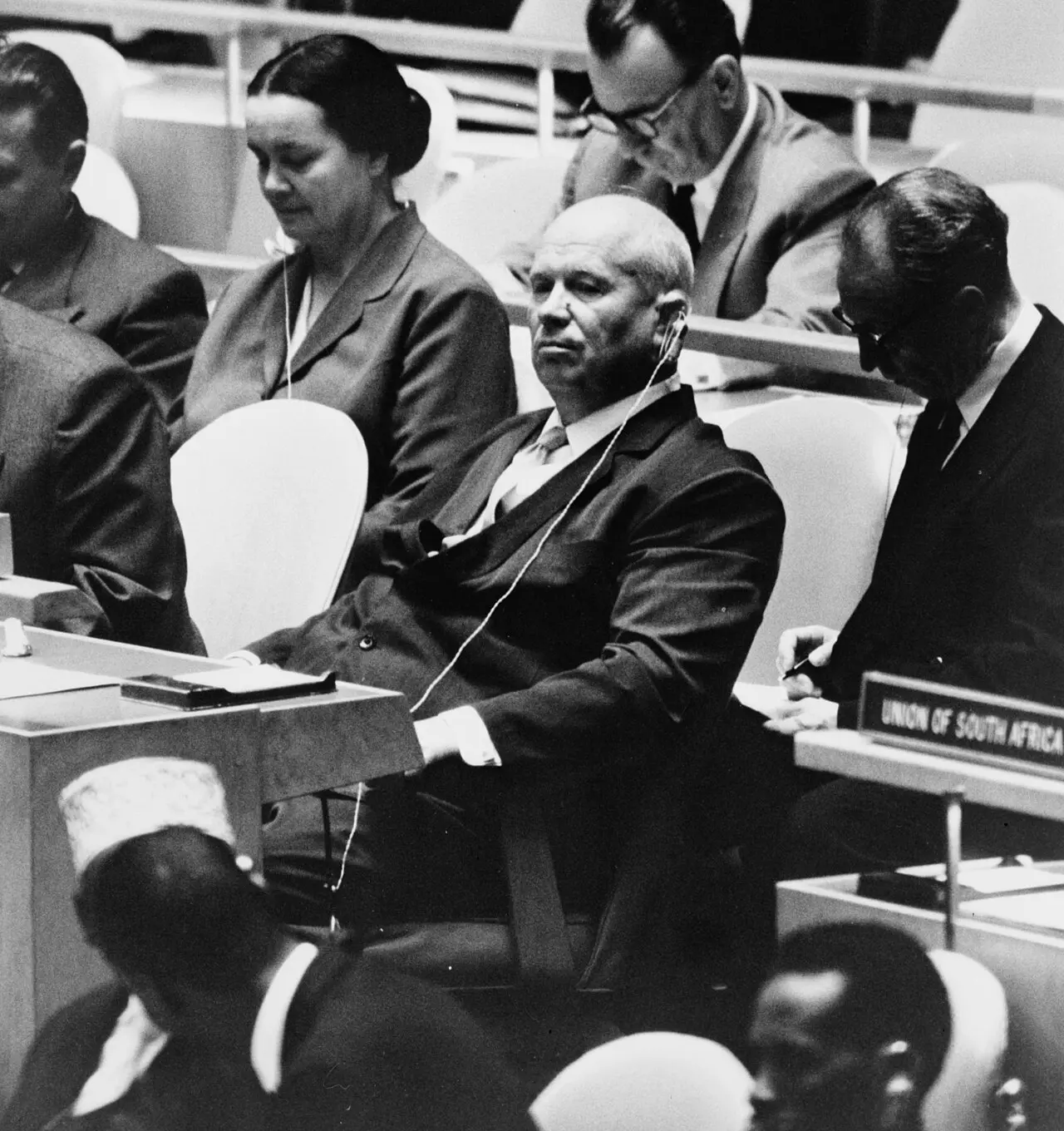 A black-and-white 1960 photograph of Soviet leader Nikita Khrushchev seated at his delegate's desk in the United Nations General Assembly hall in New York, wearing an earpiece for the translation feed.