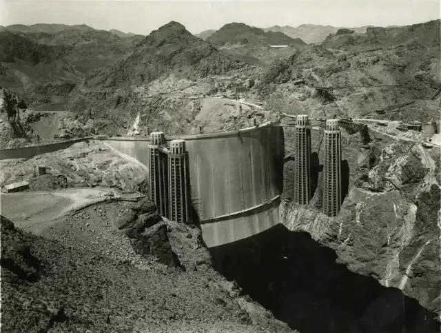A 1935 black-and-white photograph of Hoover Dam's curved concrete face with its intake towers, set in the Black Canyon of the Colorado River