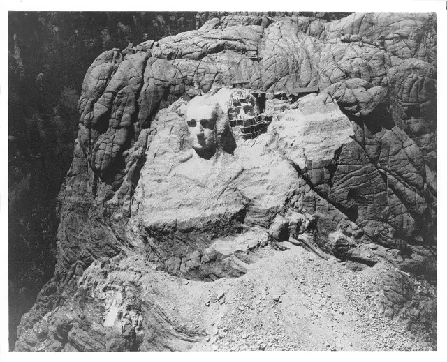 Black-and-white aerial photograph of Mount Rushmore during construction, with scaffolding on the cliff and one presidential face partially carved