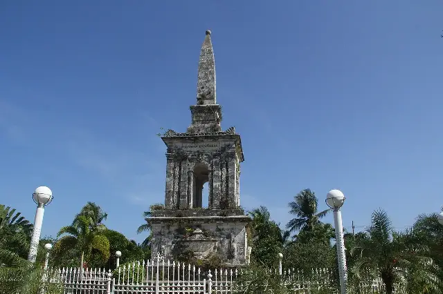 The weathered stone monument to Ferdinand Magellan at Mactan Shrine on Mactan Island, Philippines, rising above palm trees against a blue sky.