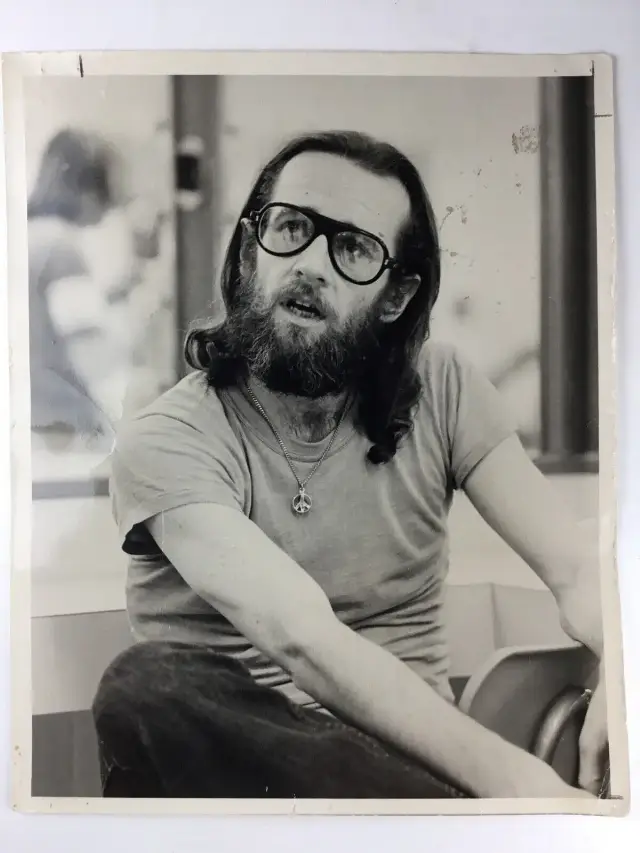A black-and-white publicity portrait from the early 1970s of comedian George Carlin, bearded and wearing glasses and a peace-sign necklace, the host of the first Saturday Night Live broadcast.