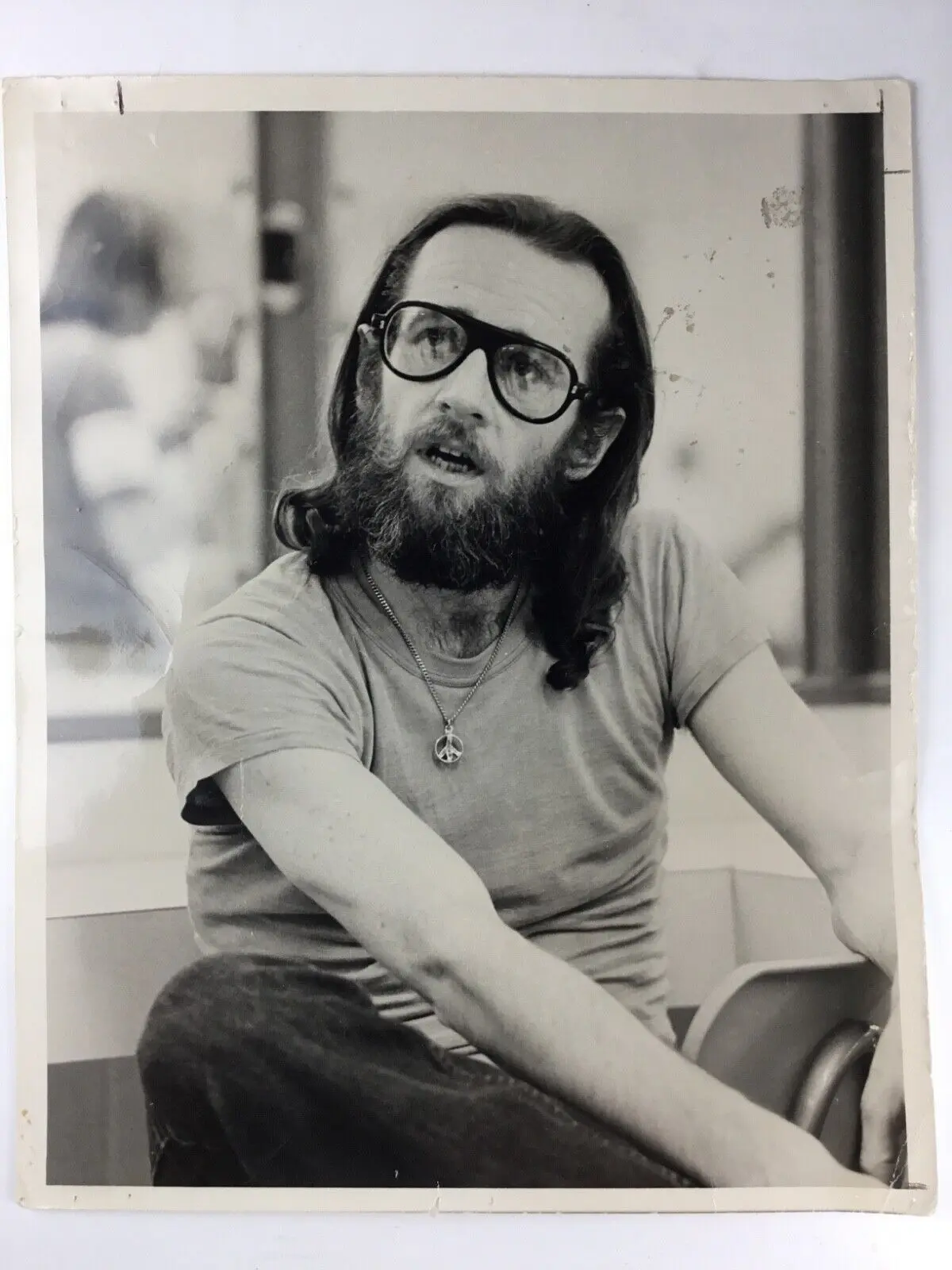 A black-and-white publicity portrait from the early 1970s of comedian George Carlin, bearded and wearing glasses and a peace-sign necklace, the host of the first Saturday Night Live broadcast.