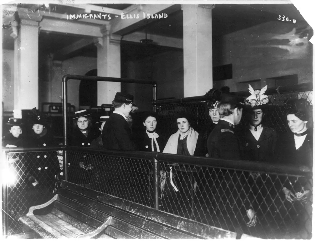 Black-and-white photograph from the early 1900s, labeled 'Immigrants - Ellis Island,' showing a group of women immigrants waiting behind railings in the station's inspection hall.