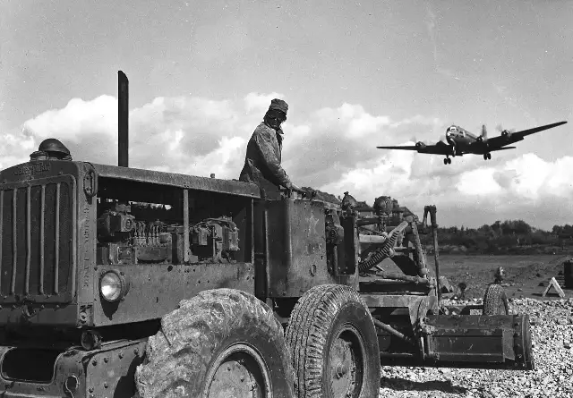 A C-54 Skymaster transport plane landing at Berlin's Tempelhof Airport during the 1948 Berlin Airlift, with a worker on a grader in the foreground.
