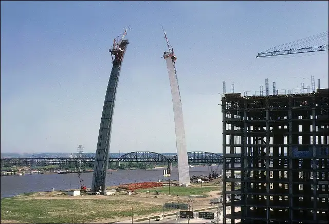 The two unfinished legs of the Gateway Arch rising over the Mississippi River in June 1965, with construction cranes perched atop each leg and the gap between them still open