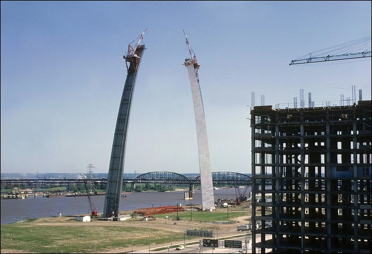 The two unfinished legs of the Gateway Arch rising over the Mississippi River in June 1965, with construction cranes perched atop each leg and the gap between them still open