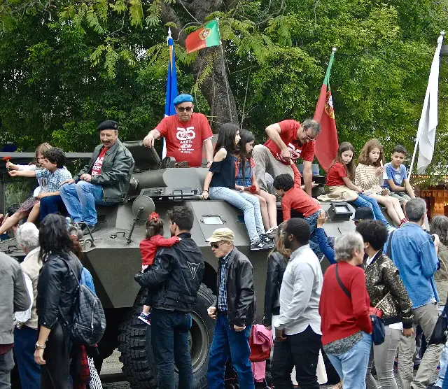 At a 2017 anniversary commemoration in Lisbon, children sit atop a Chaimite armored vehicle of the type used by rebel troops during the 1974 Carnation Revolution; marchers wear '25 Abril' shirts and a child holds a red carnation. (Photo from 2017, not 197