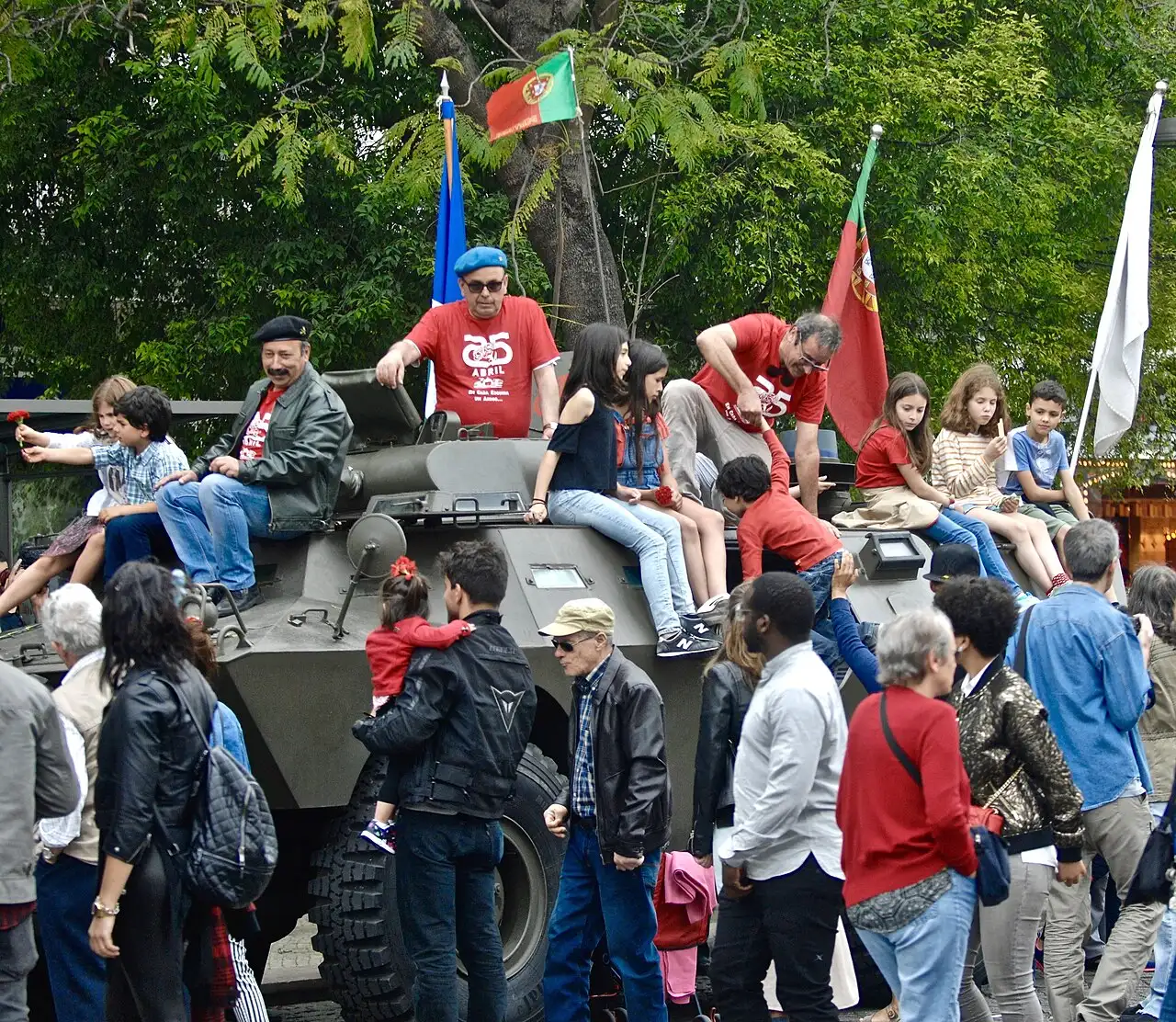 At a 2017 anniversary commemoration in Lisbon, children sit atop a Chaimite armored vehicle of the type used by rebel troops during the 1974 Carnation Revolution; marchers wear '25 Abril' shirts and a child holds a red carnation. (Photo from 2017, not 197