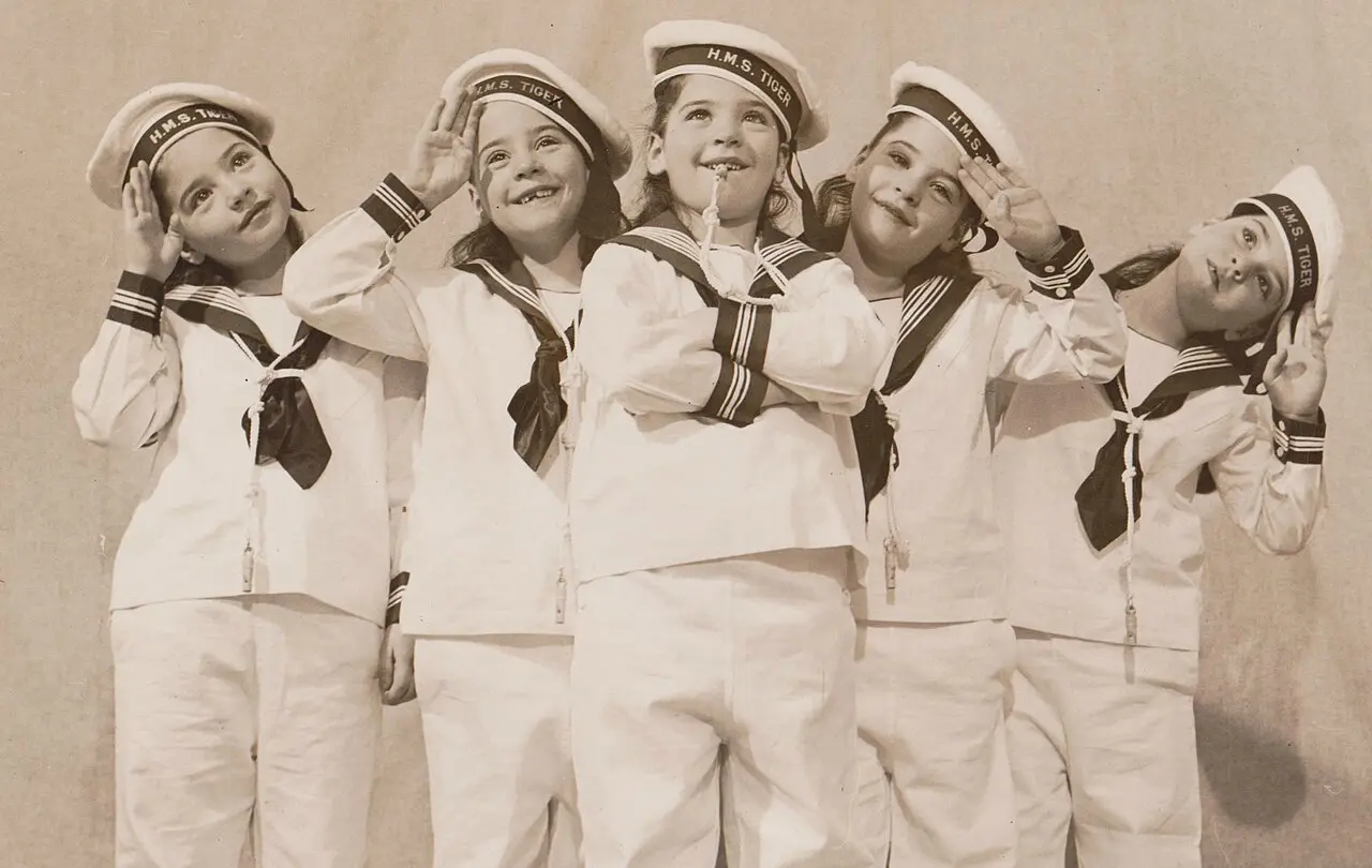 The five Dionne sisters in sailor suits and H.M.S. Tiger caps salute together in a 1930s photograph.