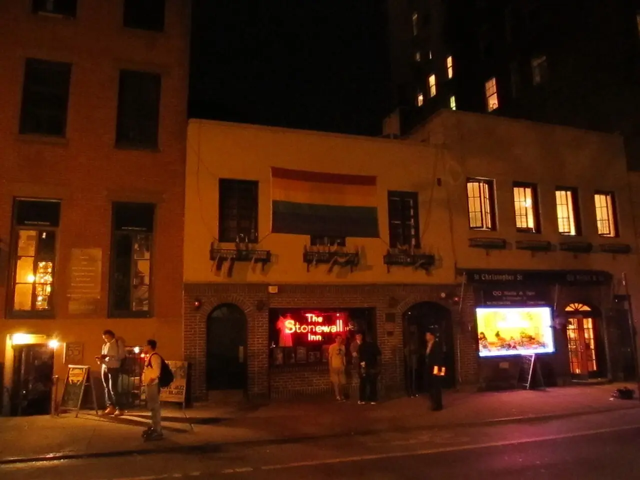 The Stonewall Inn in Greenwich Village, New York City, at night, with its neon sign and a rainbow flag above the entrance.