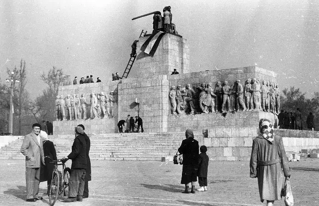 Black-and-white photograph of the empty pedestal of Stalin's toppled statue in Budapest in 1956, with people standing on it and a Hungarian flag draped over the remains.