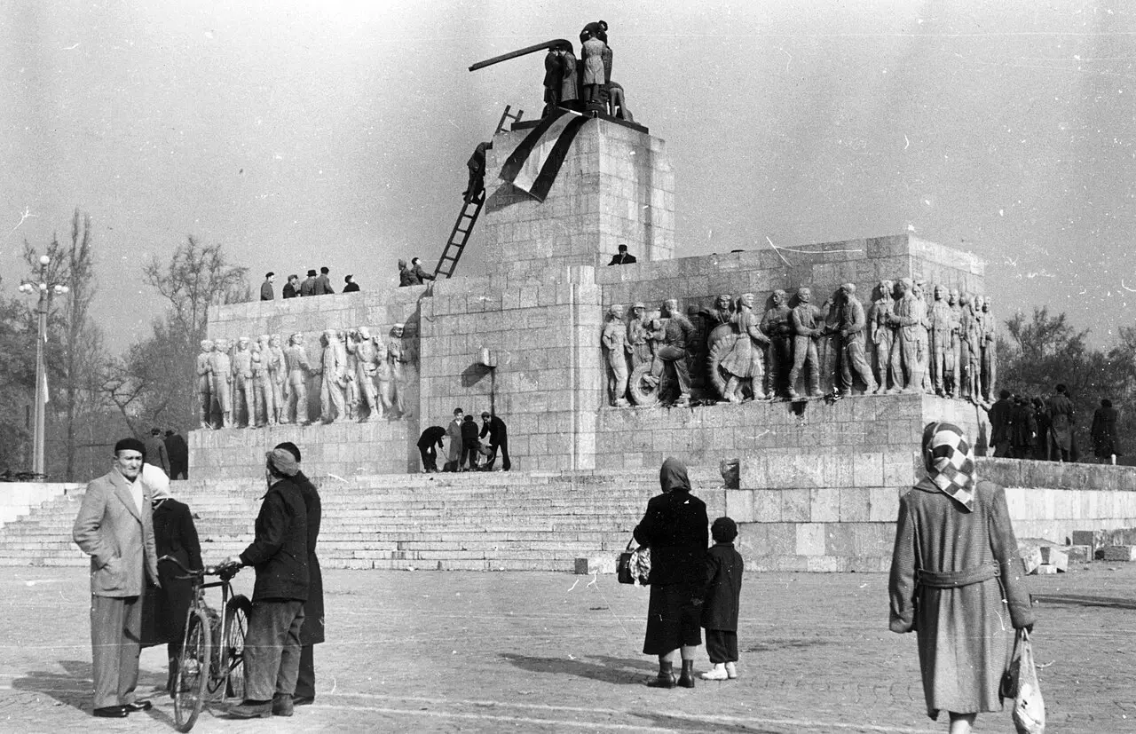 Black-and-white photograph of the empty pedestal of Stalin's toppled statue in Budapest in 1956, with people standing on it and a Hungarian flag draped over the remains.