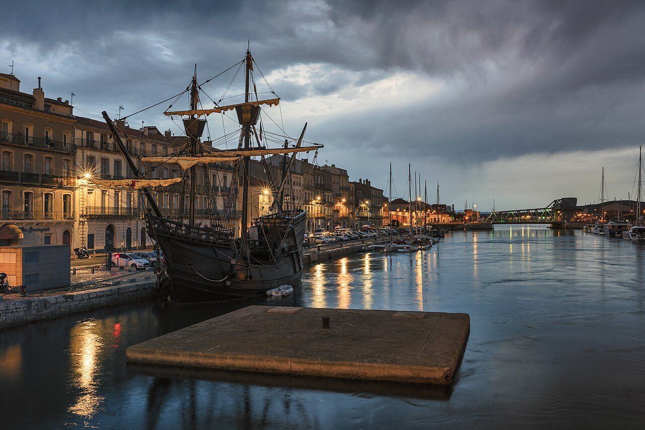 A full-size replica of the Nao Victoria, the carrack that completed the first circumnavigation of the globe, moored in a harbor at dusk.
