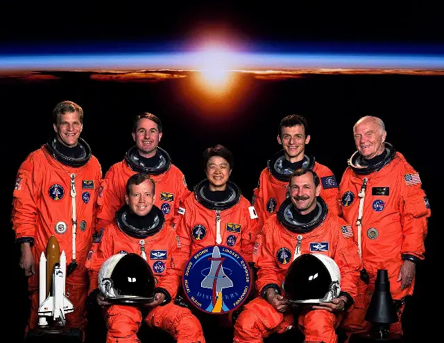 The seven-member STS-95 crew portrait: six astronauts in orange flight suits seated or standing around a seated colleague in a blue flight suit, with John Glenn standing at the far right