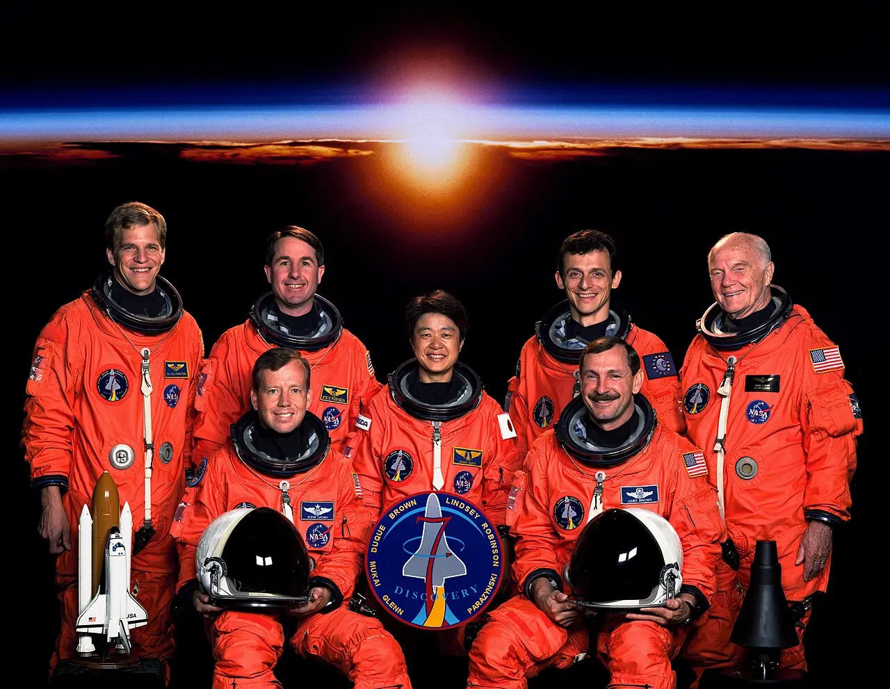 The seven-member STS-95 crew portrait: six astronauts in orange flight suits seated or standing around a seated colleague in a blue flight suit, with John Glenn standing at the far right