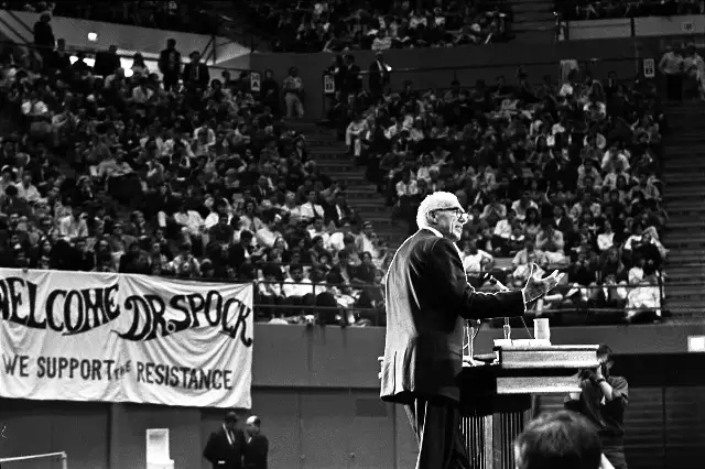 Dr. Benjamin Spock speaking at a podium before a large crowd at an anti-war rally in 1968.