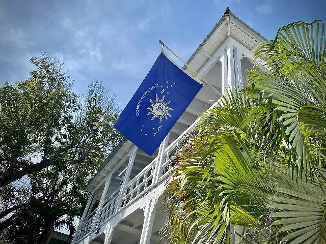 The blue flag of the Conch Republic — 'Conch Republic 1828 Key West' around a sun emblem — flying from a white wooden building among palm trees in Key West, Florida.