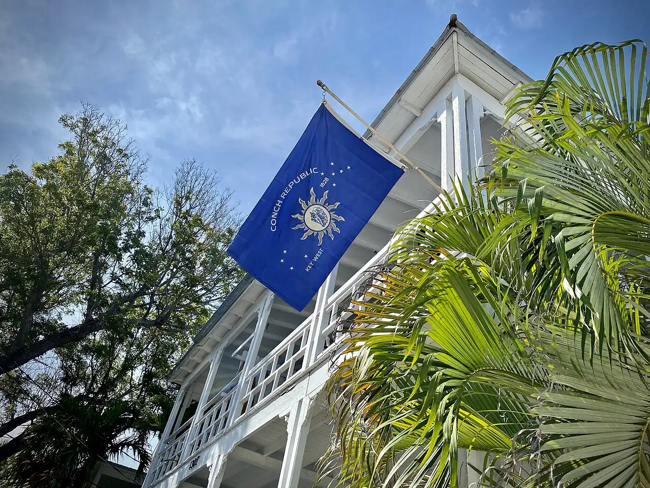 The blue flag of the Conch Republic — 'Conch Republic 1828 Key West' around a sun emblem — flying from a white wooden building among palm trees in Key West, Florida.
