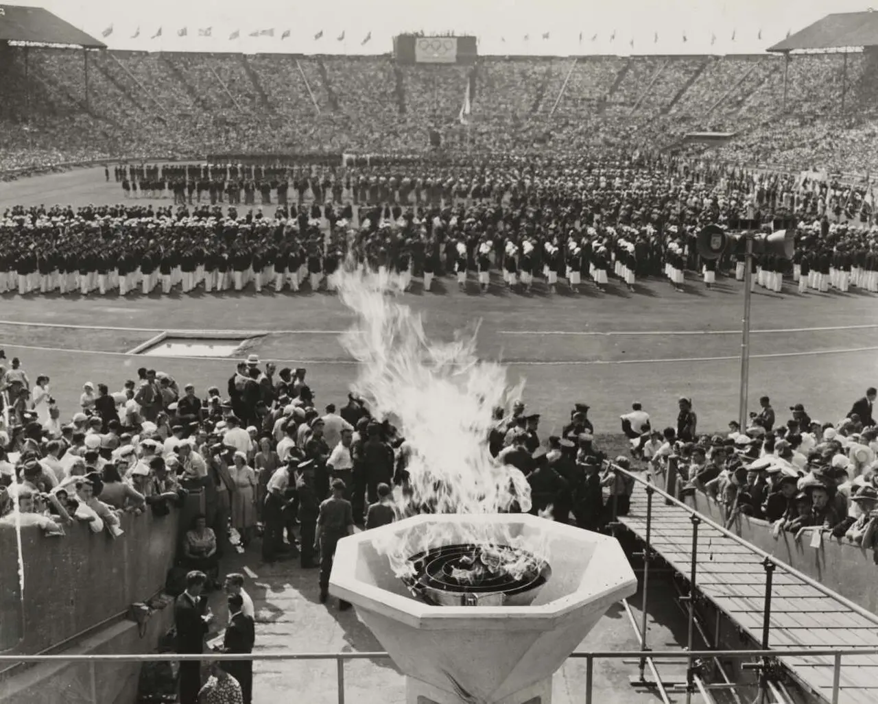 The Olympic flame burning in its bowl at Wembley Stadium during the opening of the 1948 London Games, with athletes lined up on the track and packed stands behind them.