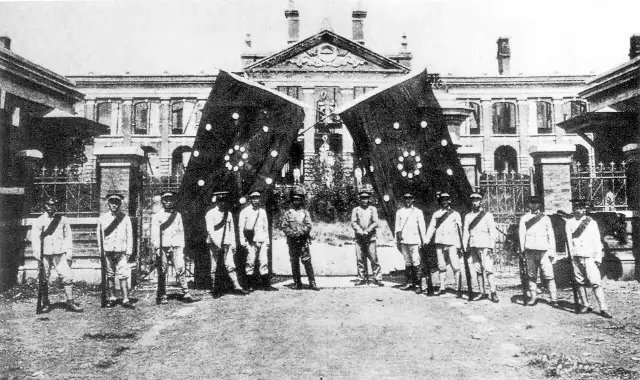 A black-and-white 1911 photograph of uniformed revolutionary soldiers holding large 18-star banners in front of the Hubei Military Government building in Wuchang.