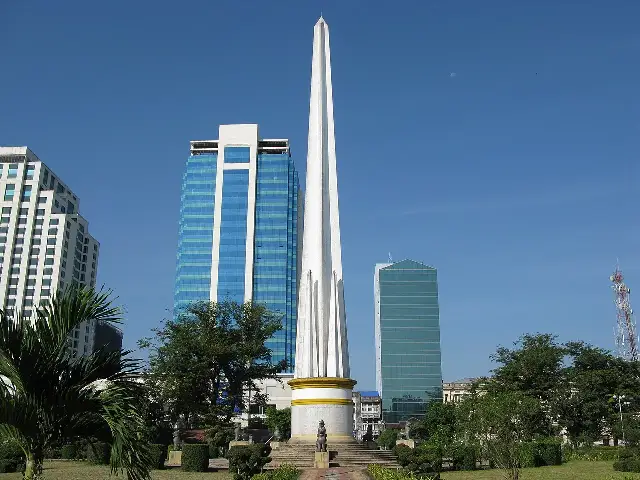 The white obelisk Independence Monument in Maha Bandula Park, Yangon, framed by palm trees against a blue sky.